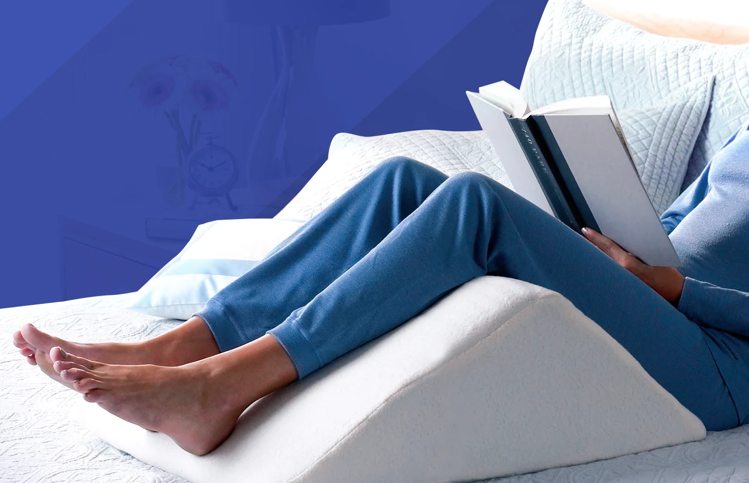 Someone in blue loungewear reads in bed with their legs raised on the white Angle wedge, white quilted bedding around them and a deep blue wall behind.