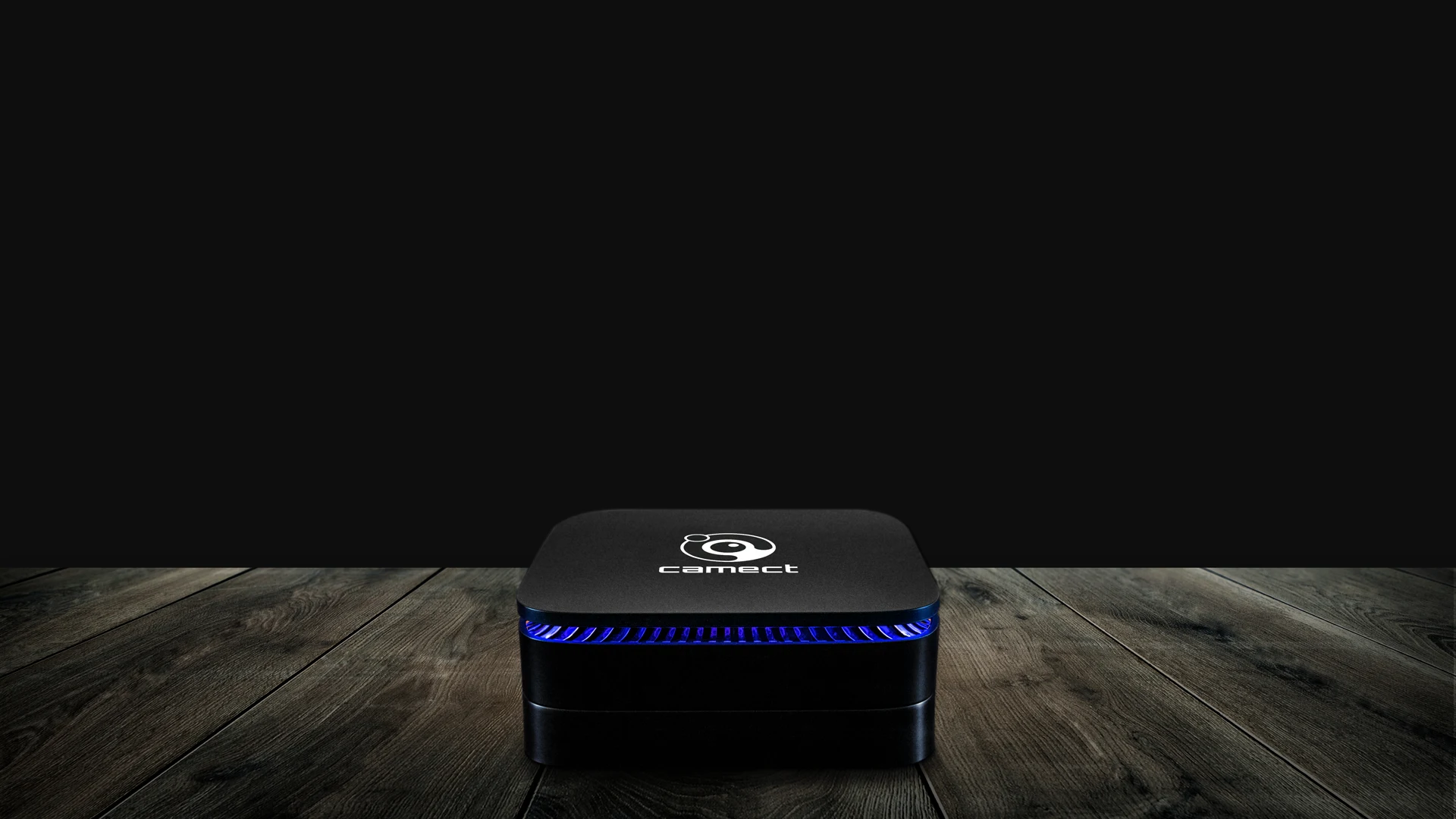 The Camect hub — a small black box with a blue light ring under its lid — sitting on dark wood planks against a black background.