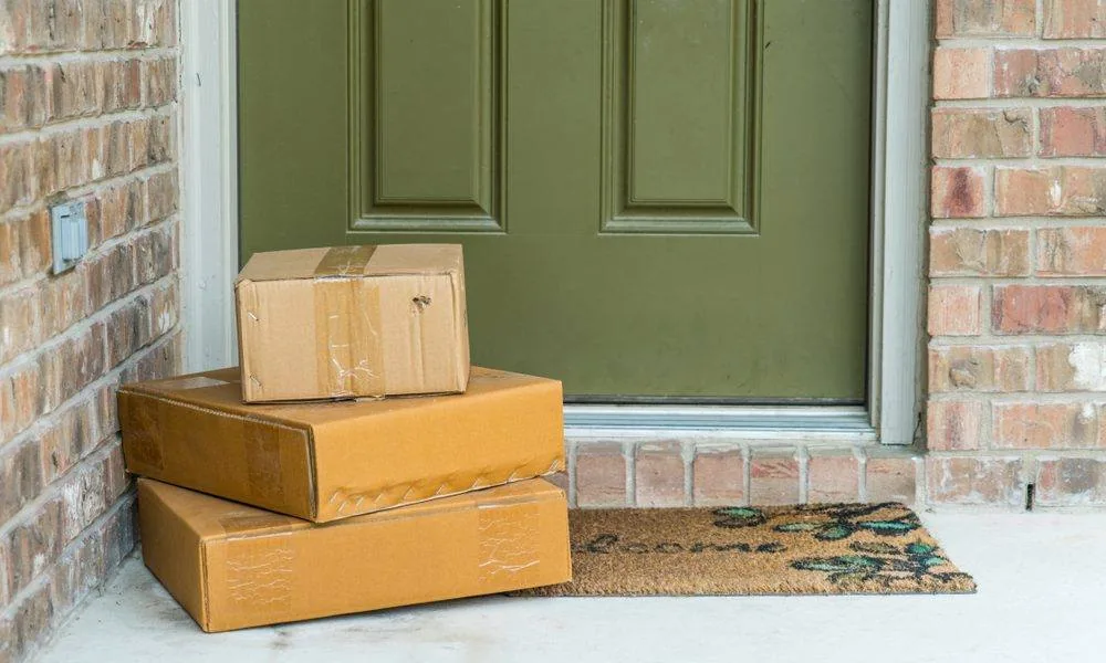 Three cardboard packages stacked by a green front door on a brick porch
