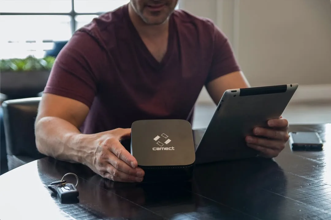 A man at a table holds a tablet beside the black Camect hub, car keys and a phone next to them
