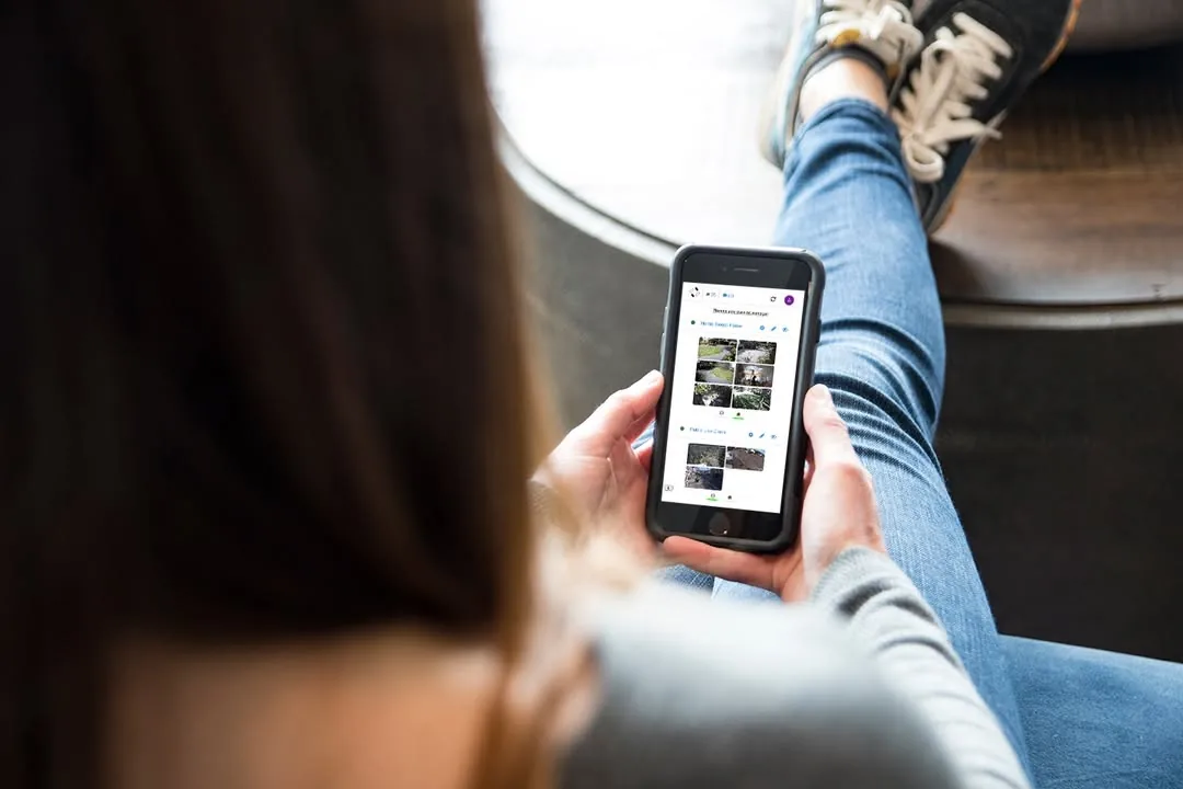 Over a woman’s shoulder: she holds a phone showing a grid of outdoor camera feeds, her jeans and sneakers propped up in front of her