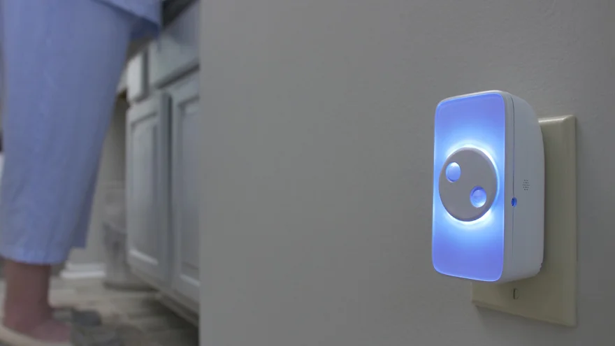 A home at night: a person’s slippered feet and pajama legs at the left edge, and the CareAlert glowing blue-white from a wall outlet on the right.