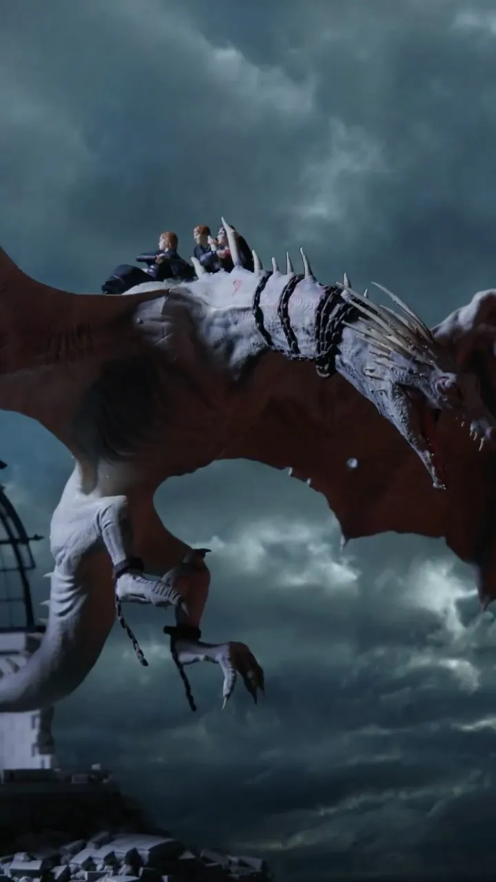 Video still: Harry, Ron and Hermione ride the back of a pale, chained dragon past a domed rooftop under storm clouds