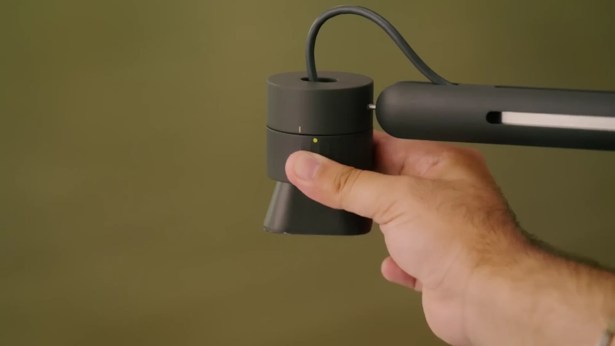 A hand holding the Logitech Reach camera head where it mounts to the end of the articulating arm, against a plain olive backdrop.