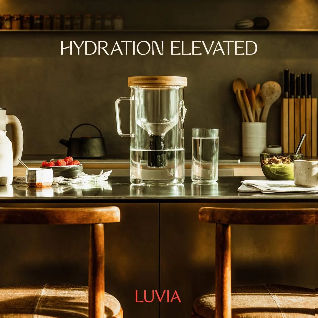 Luvia ad: the glass filter pitcher with its bamboo lid and a full glass of water on a dark kitchen counter in low golden light, raspberries and a bowl of granola beside them, two wooden stools in the foreground, under the words HYDRATION ELEVATED, with the LUVIA wordmark in red at the bottom