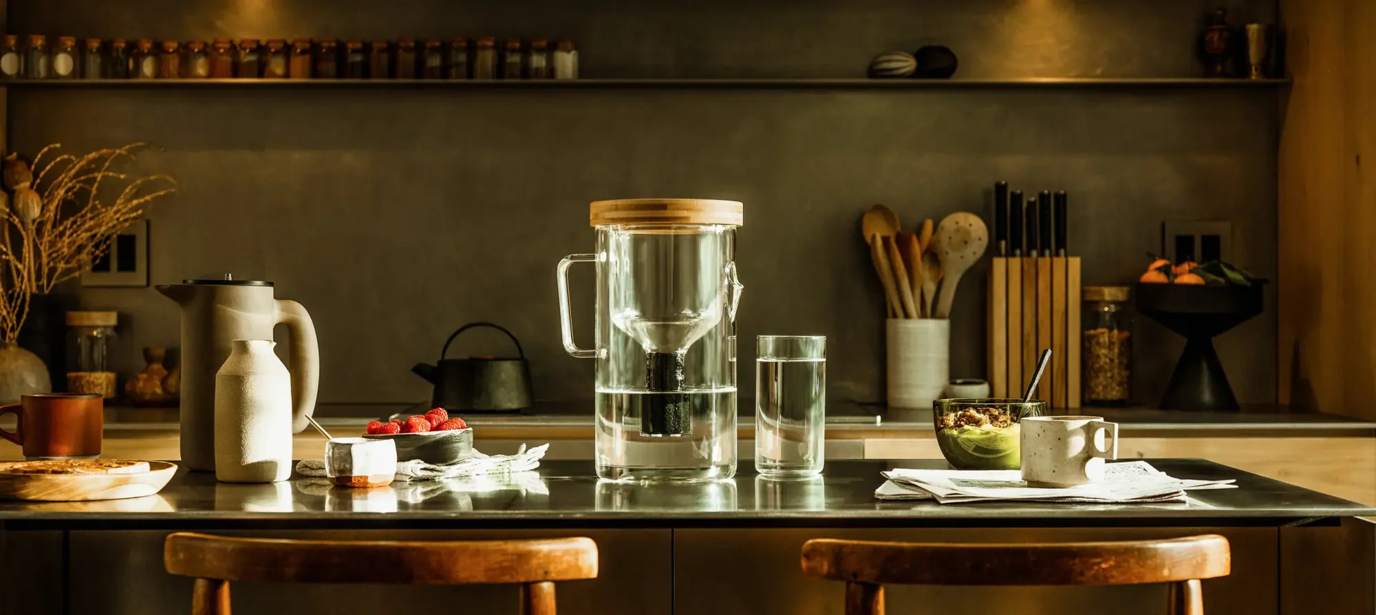 The Luvia pitcher on a dark concrete kitchen counter in warm raking light — glass body, bamboo lid, a filled glass beside it, with spice jars, a knife block and a bowl of fruit along the counter behind.