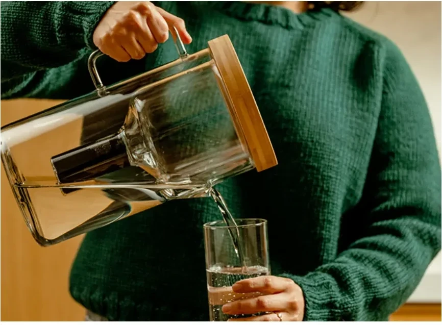 A woman in a green sweater pours water from the Luvia pitcher into a tall glass.