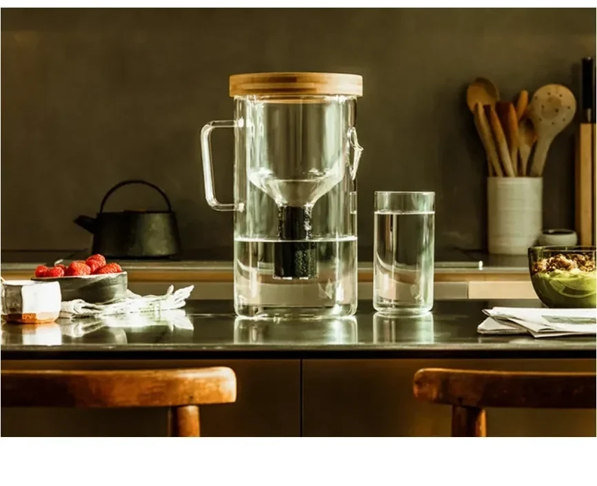 The pitcher and a filled glass on a dark kitchen counter, a cast-iron teapot and a bowl of strawberries beside them.