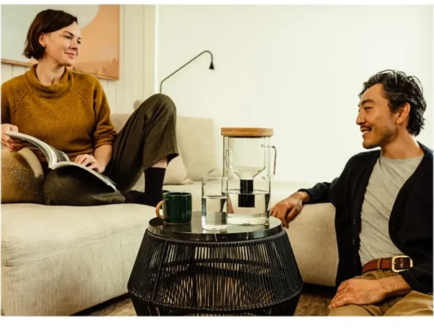 A couple relaxing in a bright living room, the pitcher and a mug on the wire side table between them.