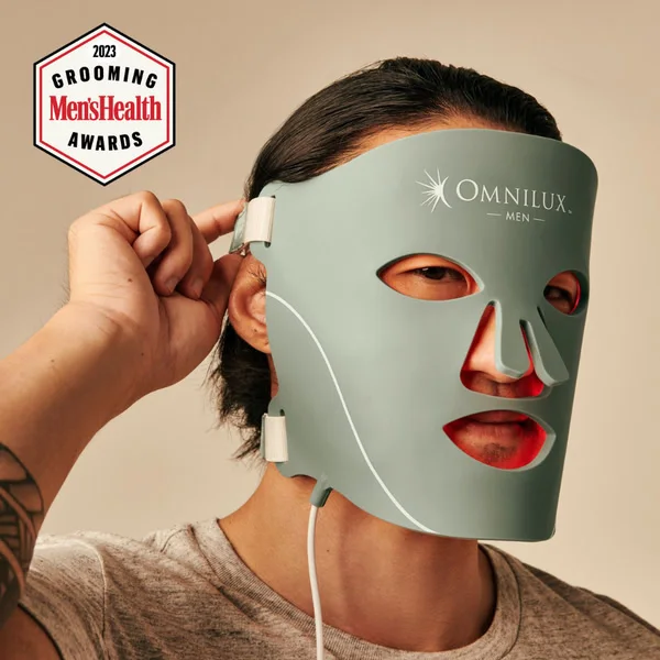 Omnilux Men ad image: a man holds the sage-green LED mask to his face, its red light showing through the eye, nose and mouth openings, under a 2023 Men’s Health Grooming Awards badge.