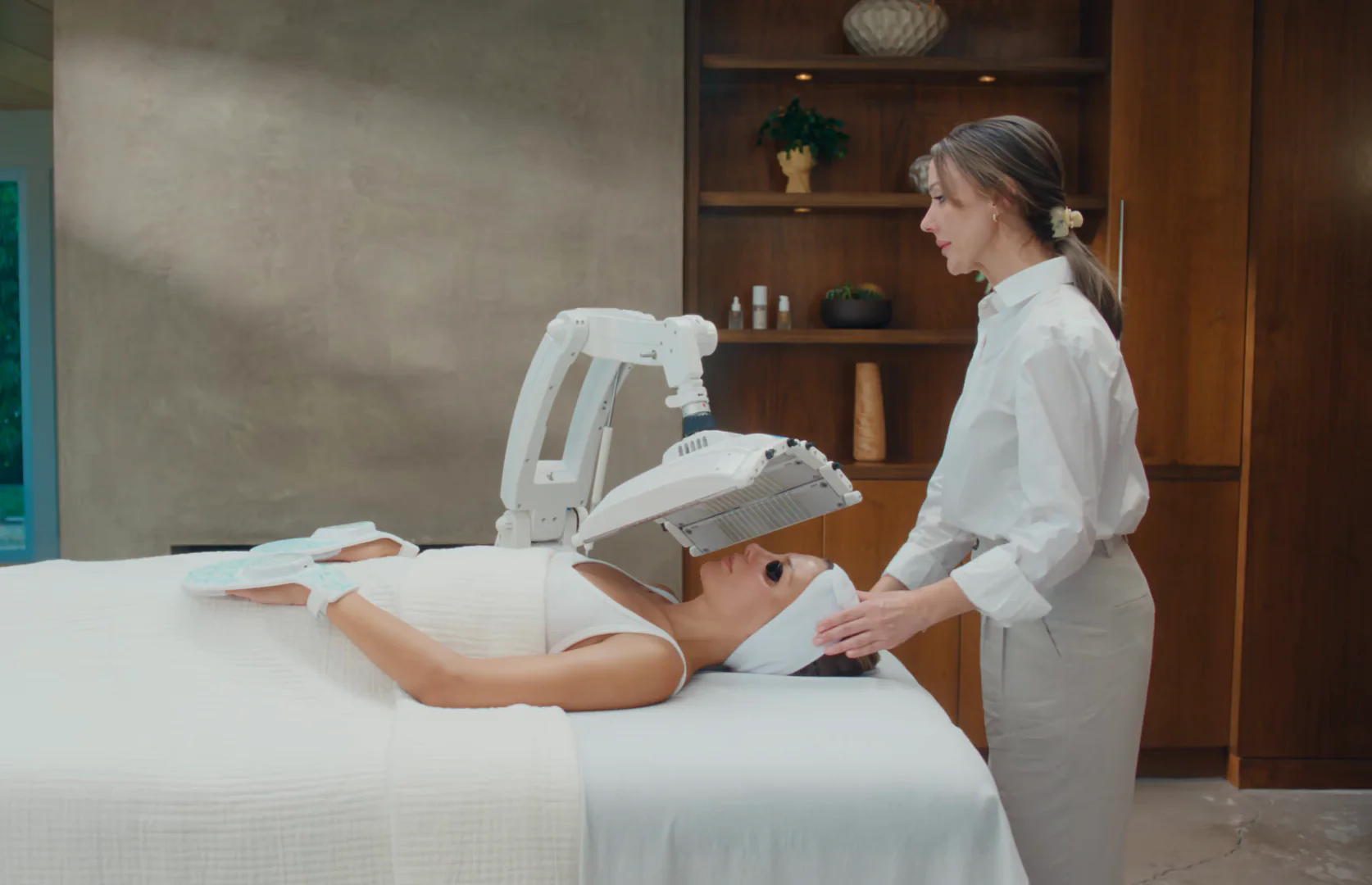 A practitioner in professional whites stands at the head of a treatment bed in a wood-panelled clinic room, positioning an articulated Omnilux LED panel over a client’s face while the flexible LED hand devices rest on the client’s hands.