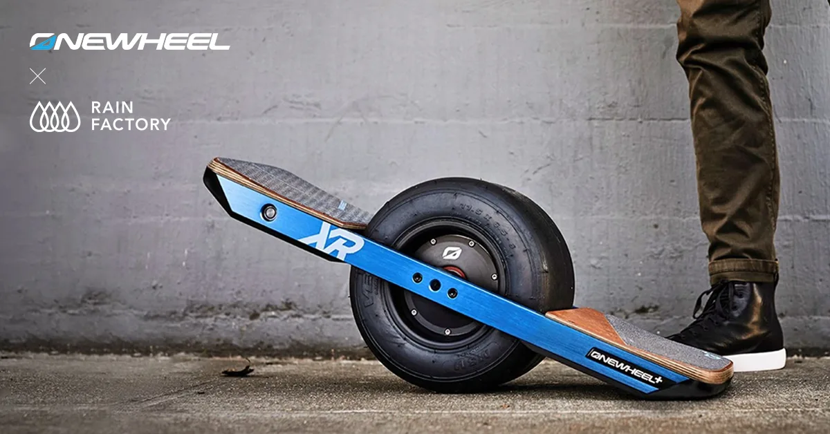 A Onewheel+ XR resting on its single fat tire on a concrete floor, tipped so one grip-taped footpad lifts clear and the other settles beside a person's black sneaker. Blue anodized rails over a wooden deck, XR lettering on the near rail, a bare concrete wall behind. An ONEWHEEL and Rain Factory lockup sits in the upper left.