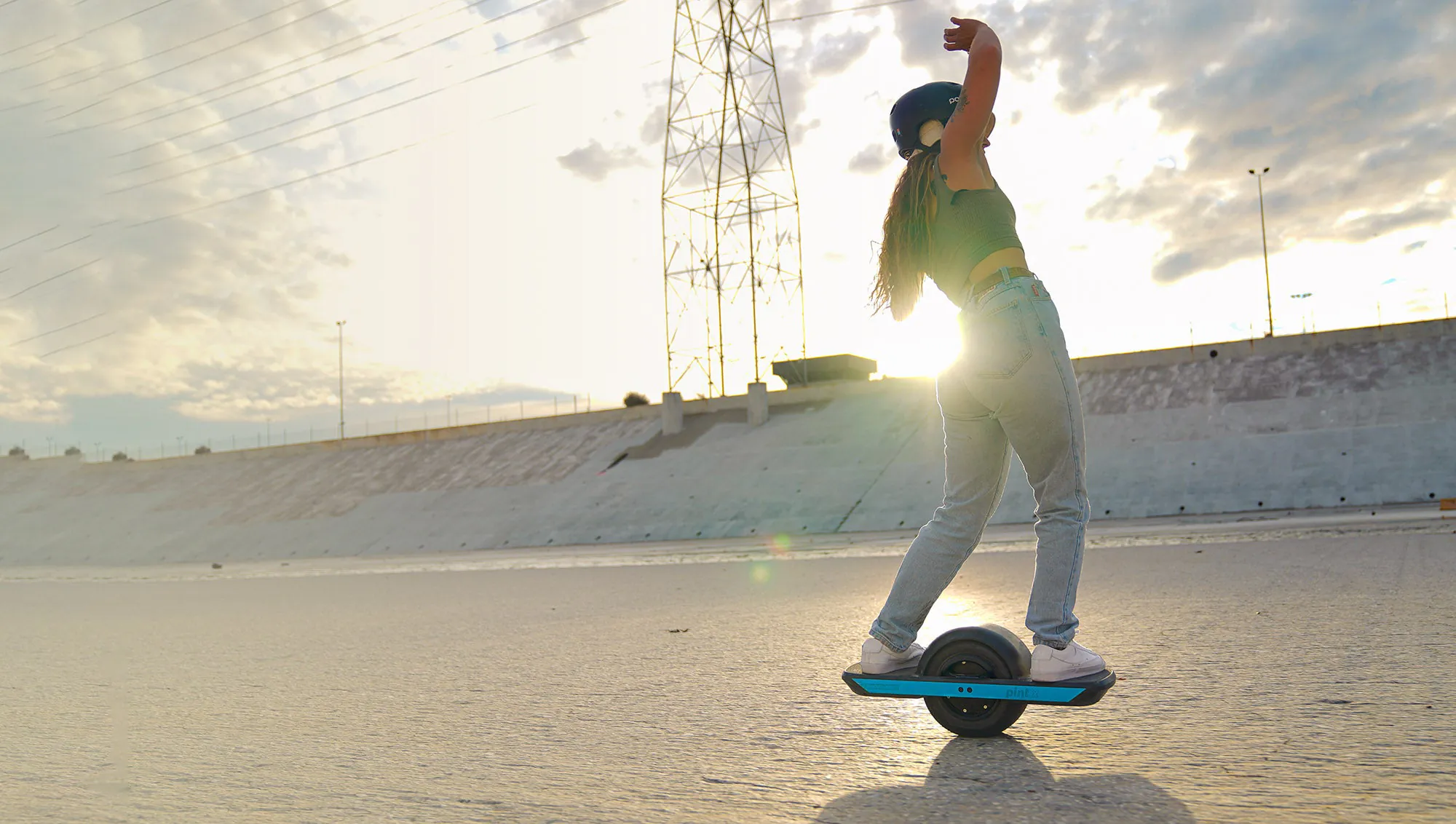 A helmeted rider carves a Pint X along the concrete floor of the Los Angeles river channel at sunset, one arm raised, the board's blue rail reading “pint X” and the sun flaring low behind a transmission tower.