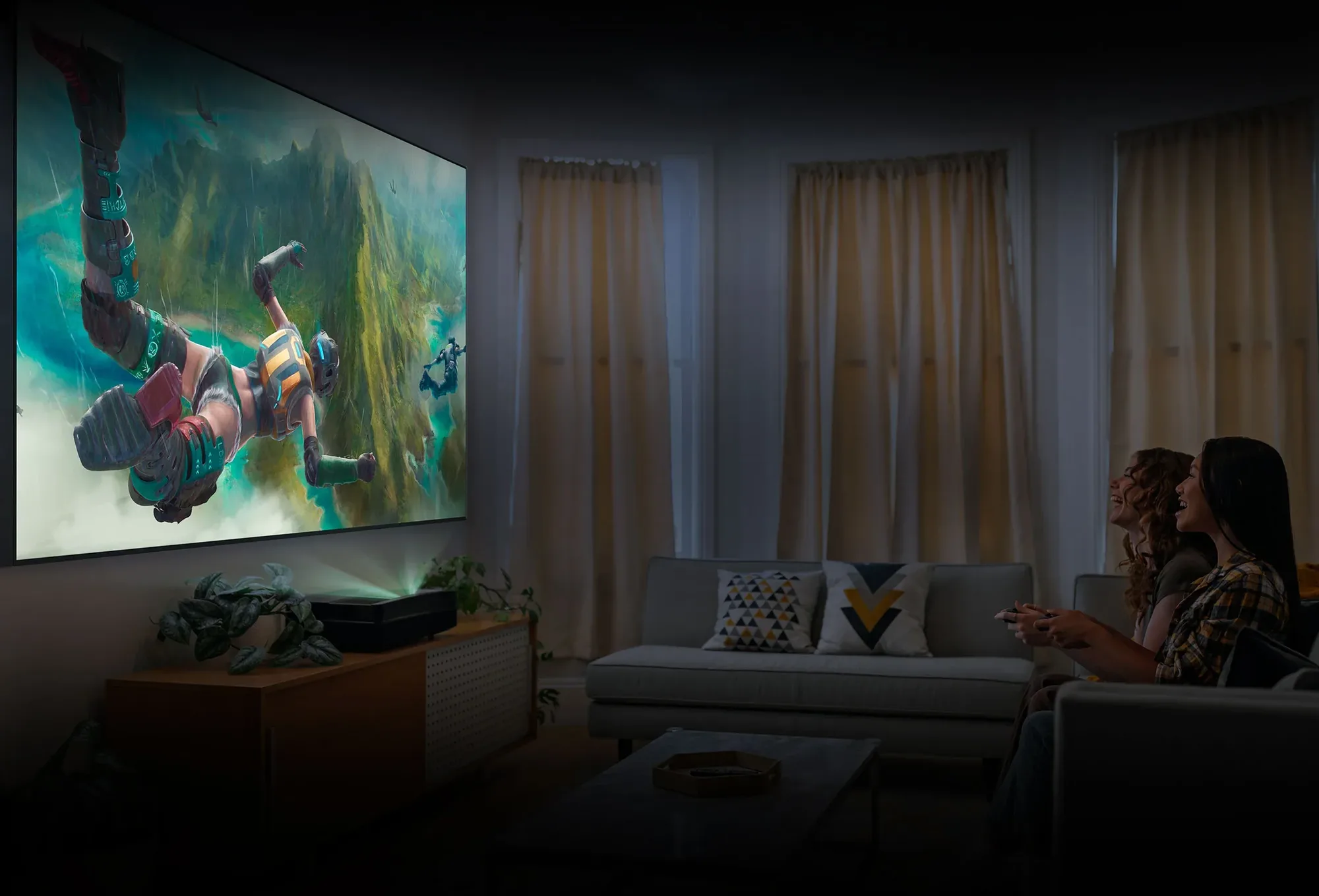 An evening living room lit only by the projected picture: two friends on a side sofa laughing over a game controller, the wall-filling game scene thrown from the projector on the cabinet beneath it.