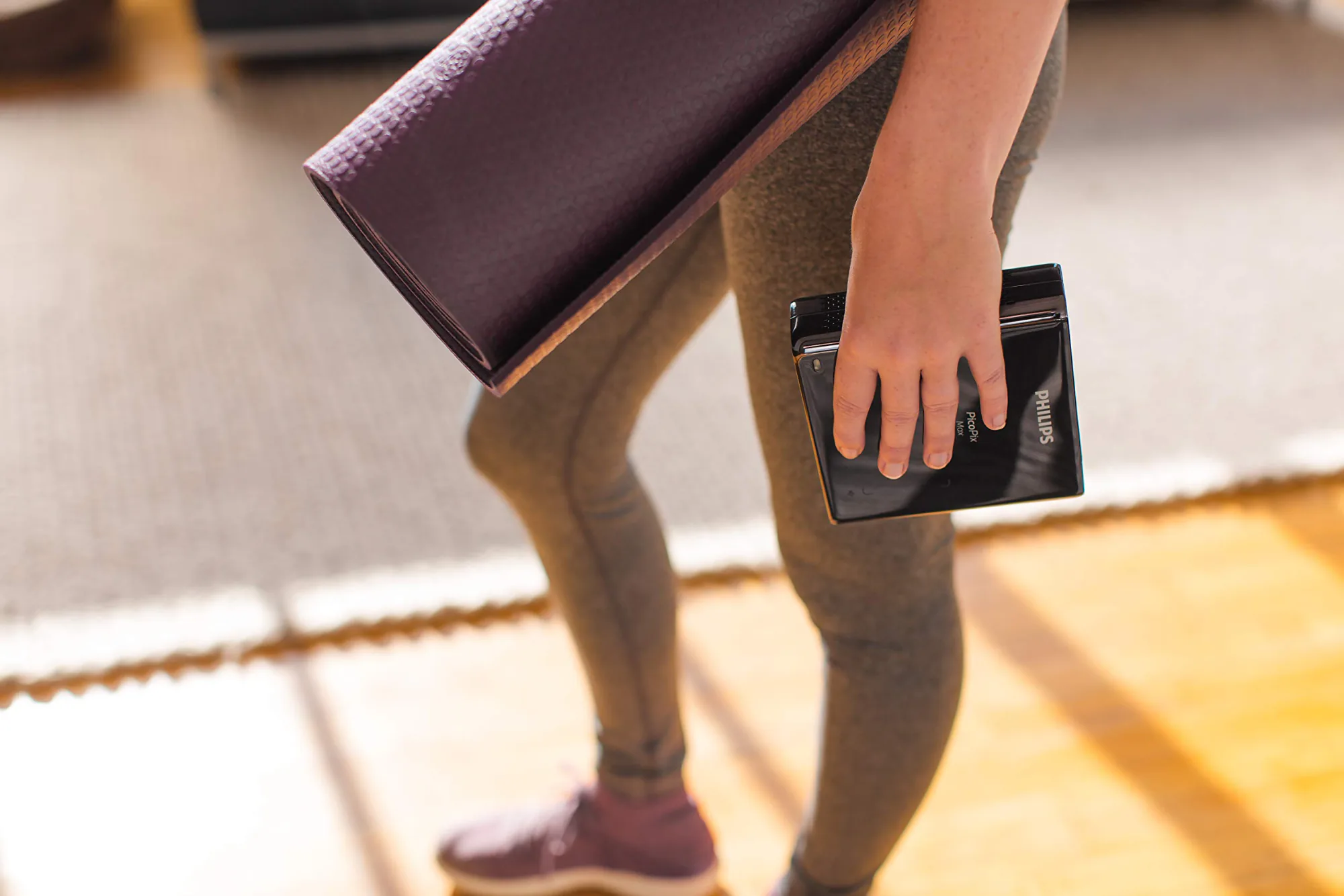 A woman crosses a sunlit room with a rolled yoga mat under her arm, the same hand carrying the PicoPix Max against her leg, the PHILIPS and PicoPix Max marks legible on its glossy black body.