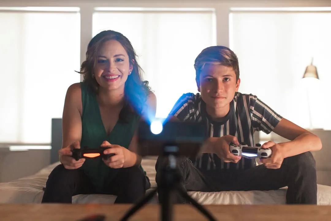 A woman and a teenage boy on a bed hold game controllers and smile, a PicoPix Max on a mini tripod in front of them, its lens lit