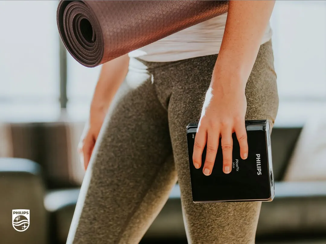 A woman in leggings carries a rolled yoga mat under one arm and the PicoPix Max in the same hand