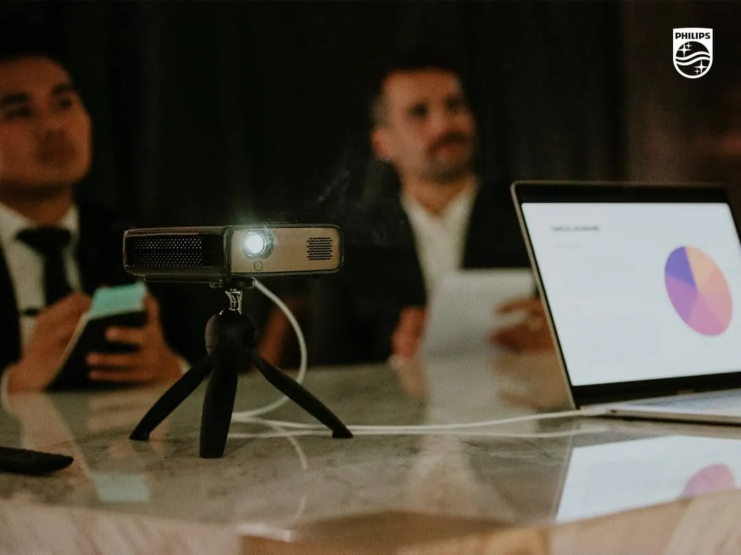 The PicoPix Max on a mini tripod on a conference table, its lens lit, beside a laptop showing a pie chart, two men in suits blurred behind it