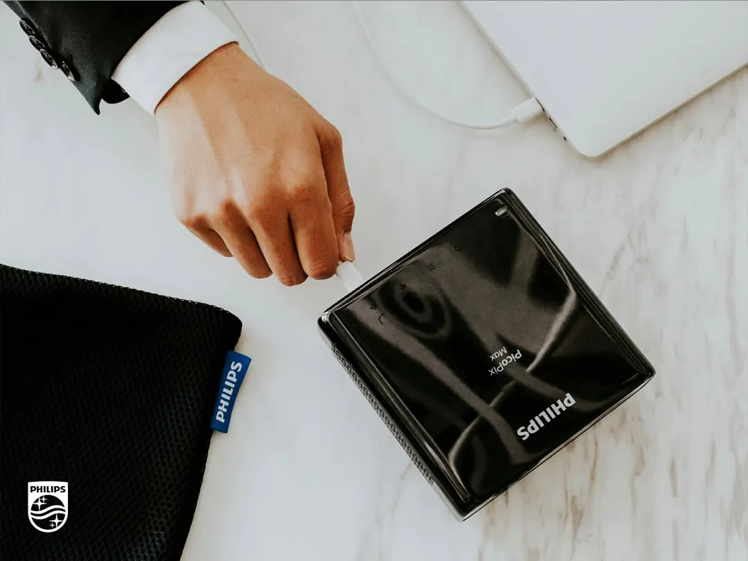 Overhead: a hand in a suit sleeve plugs a cable into the black PicoPix Max on a marble table, beside a Philips pouch and a laptop