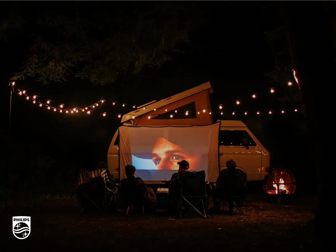 At night, people in camp chairs watch a film projected onto a sheet hung from a pop-top camper van, string lights overhead and a fire pit glowing at right
