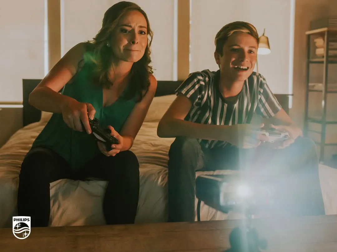 A woman and a teenage boy on a bed play a video game with controllers, the projector’s light flaring in the foreground