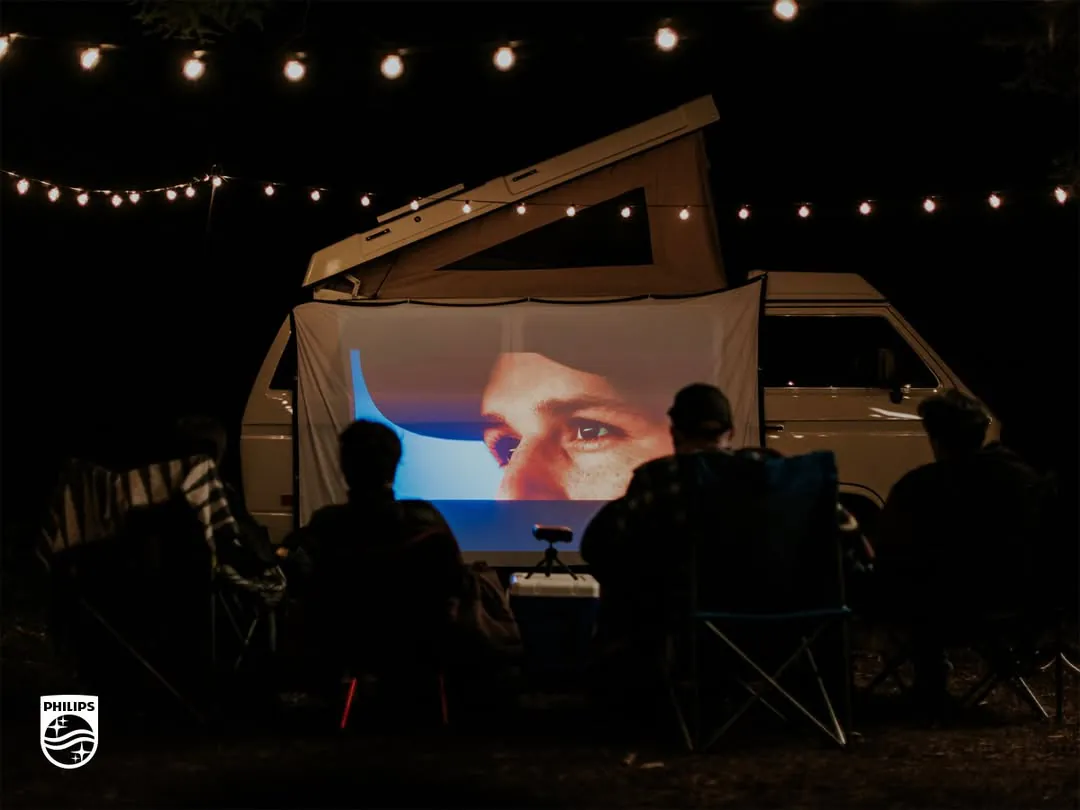 At night, a group in camp chairs watches a close-up of a face projected on a sheet hung from a camper van, the PicoPix Max set on a cooler in front of it, string lights overhead