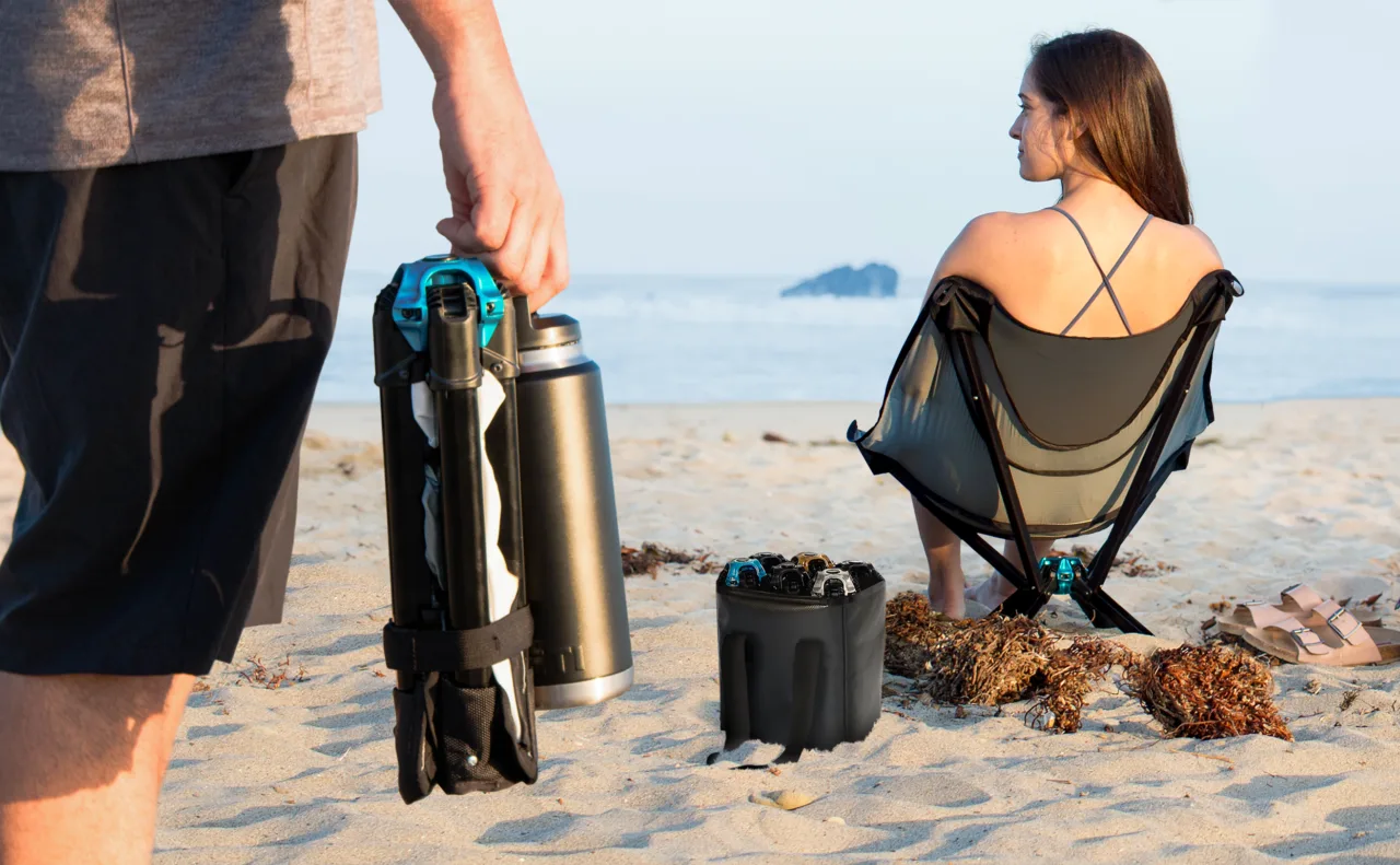 On a beach, a man carries a folded CLIQ chair and a bottle while a woman sits in an open CLIQ chair facing the water.