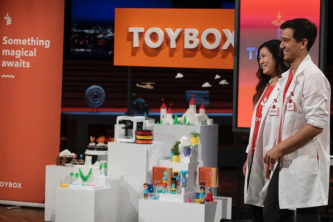 Toybox founders Jenn Chin and Ben Baltes in white lab coats on the Shark Tank set, beside white pedestals of 3D-printed toys, a castle and a small white printer, under an orange TOYBOX sign.