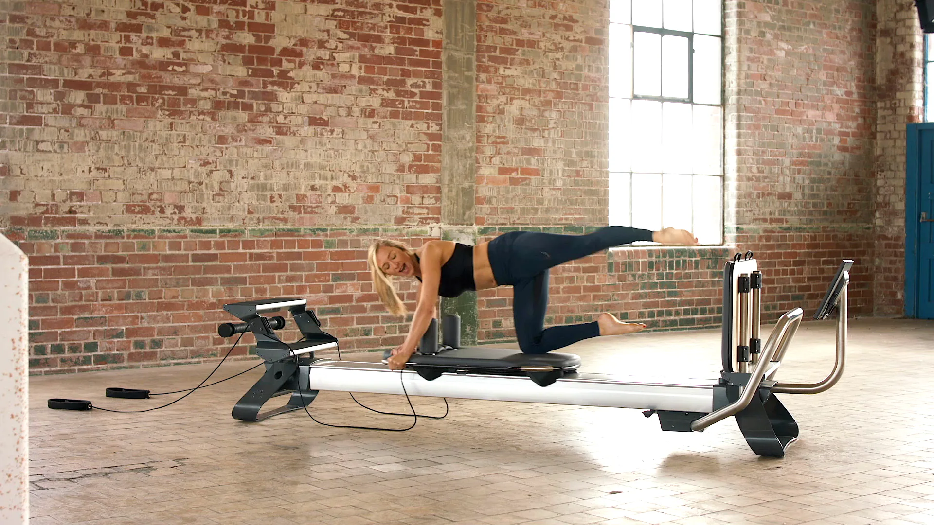 A woman works out on the Reform RX reformer — silver rail, black carriage and a touchscreen mounted at the foot end — in a raw brick loft studio, hard daylight coming through a factory window behind her.
