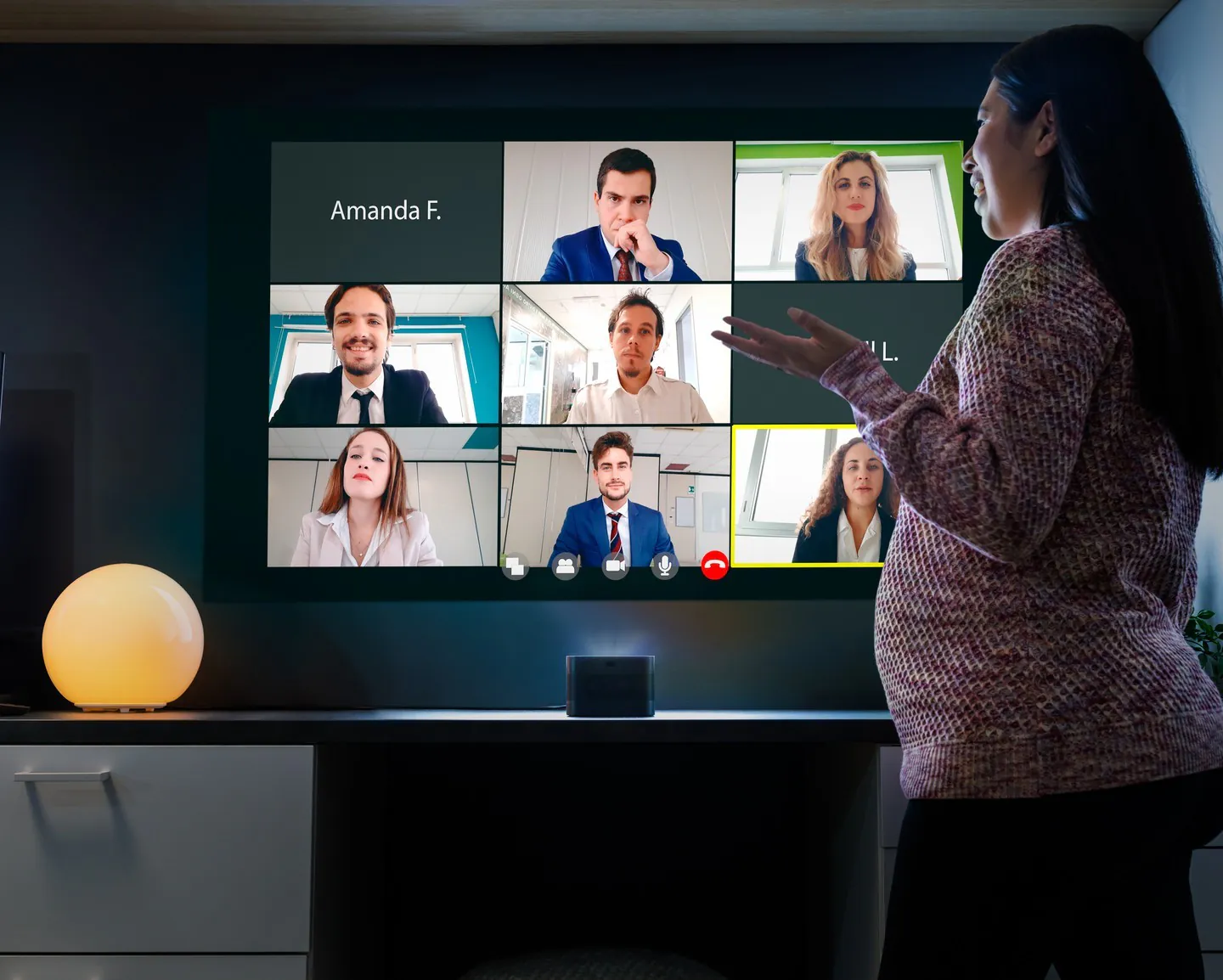 A woman gestures at a grid of video-call participants projected on the wall above a Screeneo U4 on a low cabinet