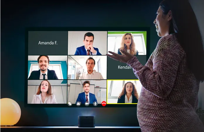 A woman gestures at a nine-person video call projected on a dark wall, the U4 on the sideboard beneath it.