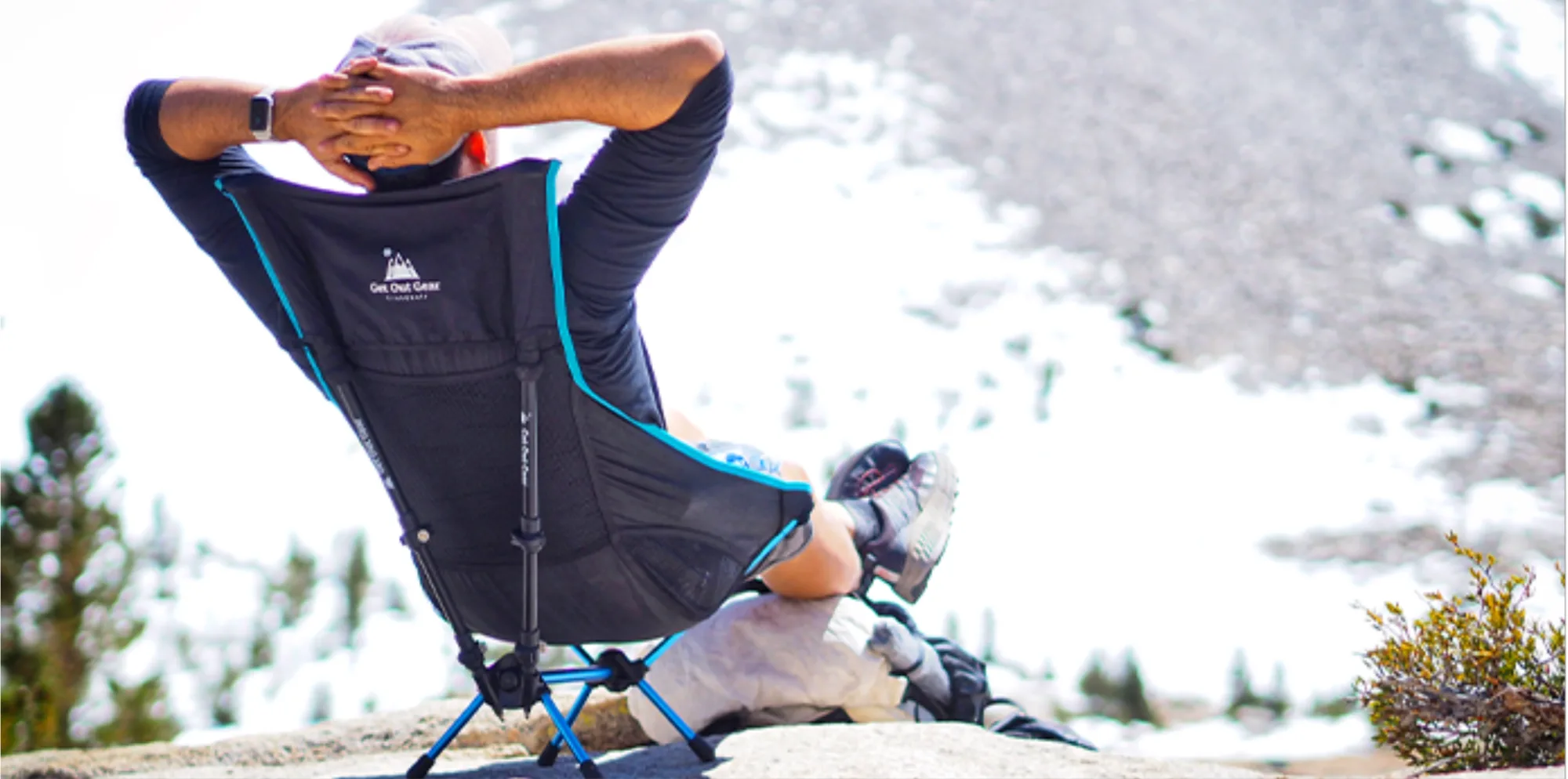 Hiker reclining in the TrekChair on a granite outcrop with arms crossed behind the head, facing a snowfield — the chair's blue-trimmed frame planted in the rock.